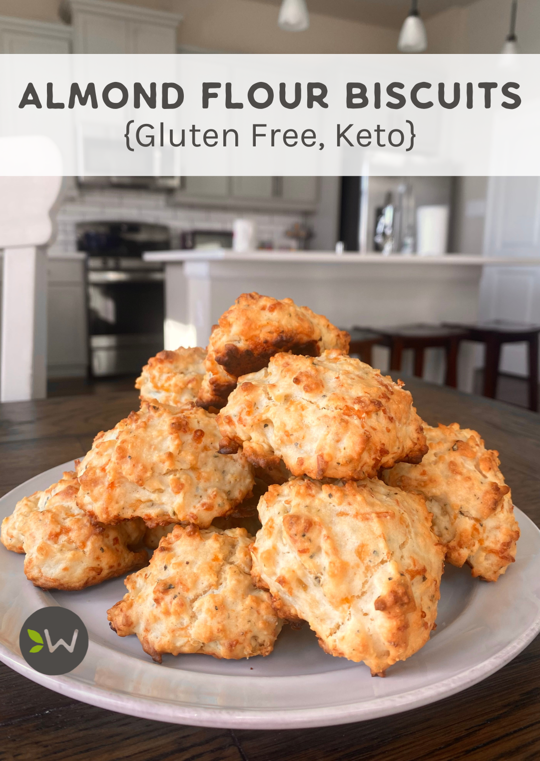 Photo of almond flour drop biscuits heaped on a white plate on a table with a kitchen in the background.